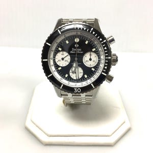 May include: A stainless steel Zodiac Sea-Chron watch with a black dial and bezel. The watch features white subdials, a tachymeter scale, and the number 30 on the bezel. The watch is displayed on a white stand.