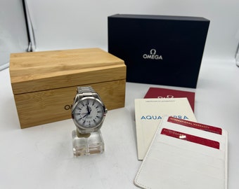 May include: A silver wristwatch with a white face and black markings. The watch is on a white display stand and is surrounded by a wooden box, a black box with the word "OMEGA" on it, and a white card with the word "AQUA" on it.