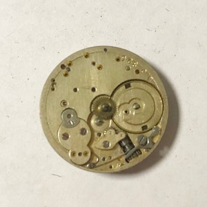 Puede incluir: Primer plano de un movimiento de reloj de tono dorado con engranajes intrincados y un dise&ntilde;o circular. El movimiento se coloca sobre un fondo blanco.