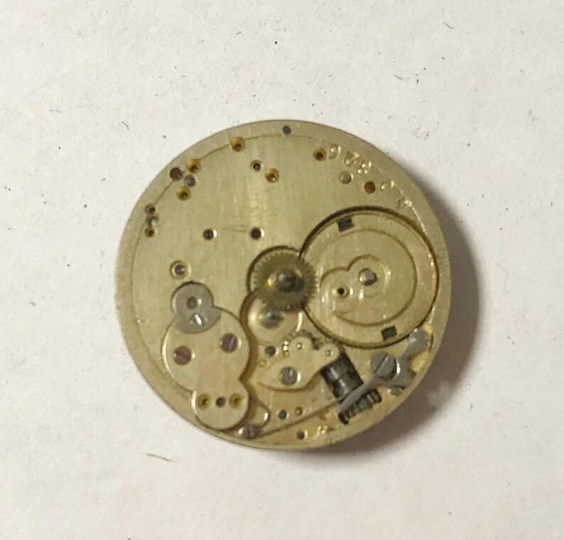 Puede incluir: Primer plano de un movimiento de reloj de tono dorado con engranajes intrincados y un dise&ntilde;o circular. El movimiento se coloca sobre un fondo blanco.