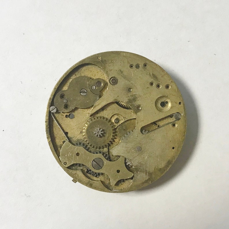Pu&ograve; includere: Primo piano di un movimento di orologio vintage, che mostra ingranaggi, molle e leve intricati. I componenti color ottone sono disposti all'interno di una cornice circolare, rivelando la complessit&agrave; meccanica dell'orologio. Il design include piccole viti e varie parti funzionali.
