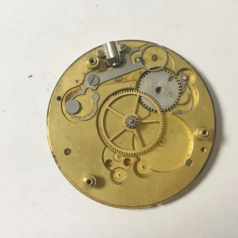 Puede incluir: Primer plano de un movimiento de reloj de color dorado con engranajes intrincados y un v&aacute;stago de cuerda de color plateado.