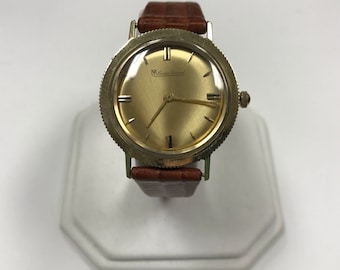 May include: A gold-toned wristwatch with a brown leather band. The watch face is round and has a gold-toned bezel. The watch face is gold and has black Roman numeral hour markers and black hands. The watch face also has the text "Le Baron Renaud" printed on it.