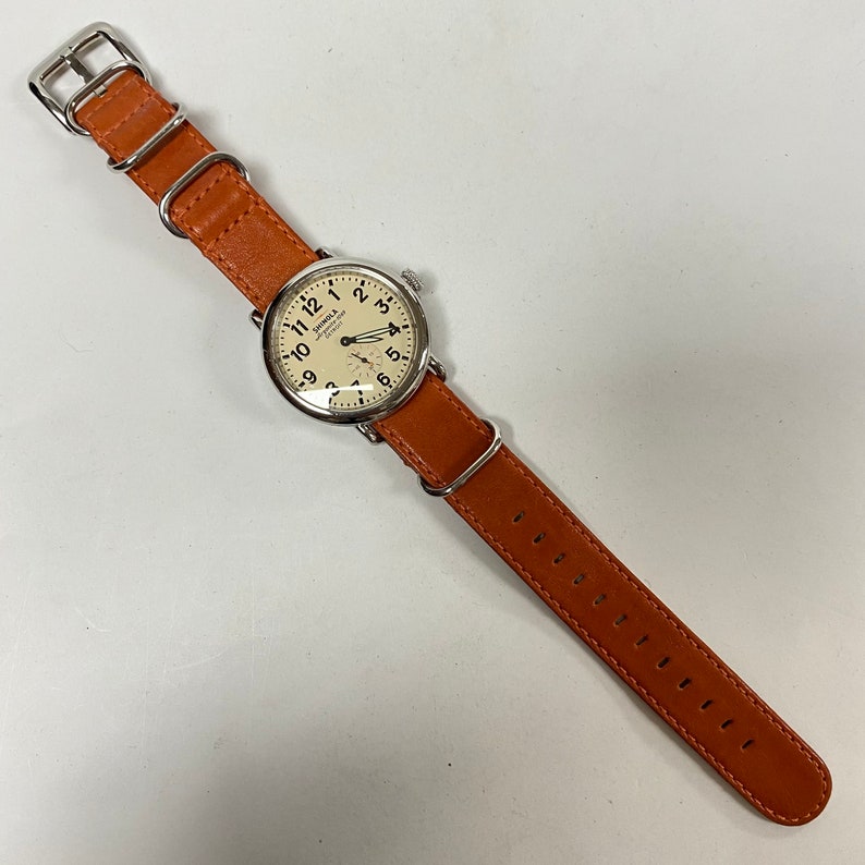 May include: A silver-toned wristwatch with a cream-colored face and black numerals. The watch has a brown leather band with a silver buckle. The watch face displays the brand name "Shinola Detroit".