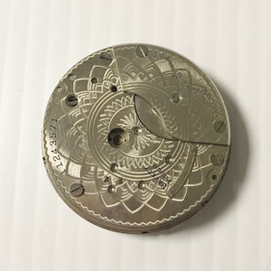 May include: A silver pocket watch movement with a decorative design and the text "Elgin Nat. Watch Co. Elgin, Ill. U.S.A." and "11243571" and "GRAVITY PINION".