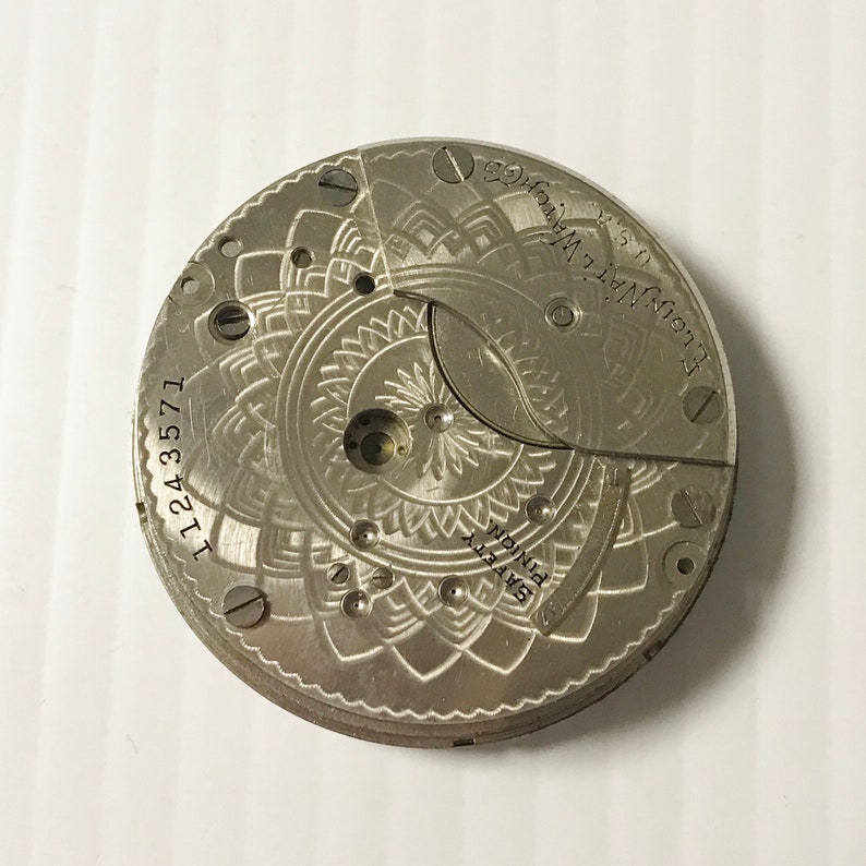 May include: A silver pocket watch movement with a decorative design and the text "Elgin Nat. Watch Co. Elgin, Ill. U.S.A." and "11243571" and "GRAVITY PINION".