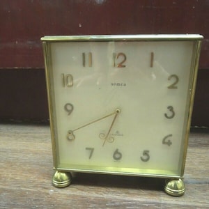 May include: A gold-toned square clock with a white face and black hands. The clock has a gold frame and gold feet. The clock face has the word "semea" printed on it and the numbers 1 through 12. The clock also has the words "8 Jewels" and "Swiss" printed on it.