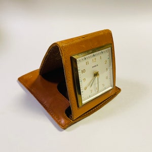 May include: A brown leather travel alarm clock with a white face and gold trim. The clock face has black numbers and hands, and the word "Seneca" printed on it.
