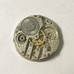 K&ouml;nnte beinhalten: Ein silbernes Uhrwerk mit freiliegenden Zahnr&auml;dern. Das Werk ist kreisf&ouml;rmig und hat einen Durchmesser von etwa 6,5 cm. Die Zahnr&auml;der sind aus Messing und in einem komplexen Muster angeordnet.