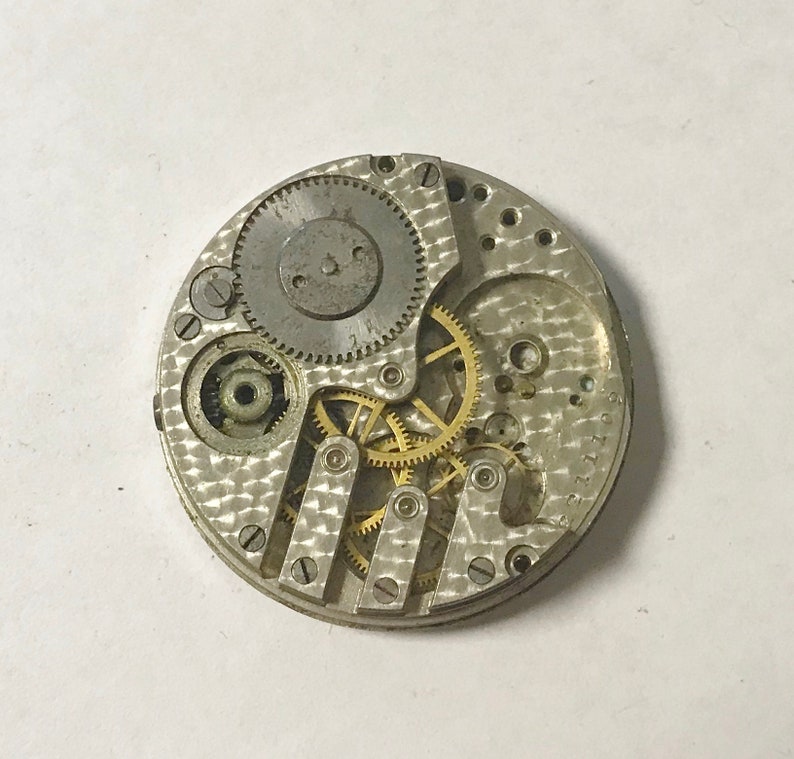K&ouml;nnte beinhalten: Ein silbernes Uhrwerk mit freiliegenden Zahnr&auml;dern. Das Werk ist kreisf&ouml;rmig und hat einen Durchmesser von etwa 6,5 cm. Die Zahnr&auml;der sind aus Messing und in einem komplexen Muster angeordnet.