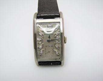 May include: A silver-toned Hamilton wristwatch with a rectangular face and diamond accents around the bezel. The watch has a black leather band.
