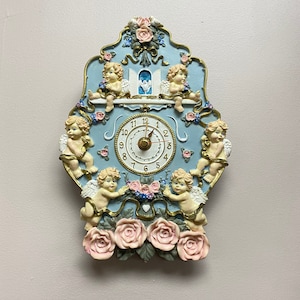 May include: A blue and white clock with cherub figurines and pink roses. The clock face has Roman numerals and a gold-colored hour and minute hand.