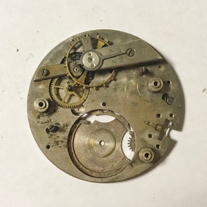 Op de afbeelding: Een close-up van een zilveren uurwerk met zichtbare tandwielen en een opwindas. Het uurwerk is gemarkeerd met "Swiss" en "R.W. 17".