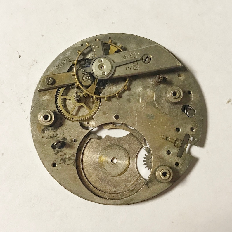 Op de afbeelding: Een close-up van een zilveren uurwerk met zichtbare tandwielen en een opwindas. Het uurwerk is gemarkeerd met "Swiss" en "R.W. 17".