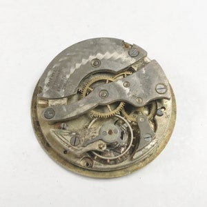 Puede incluir: Primer plano de un movimiento de reloj vintage con engranajes y componentes met&aacute;licos visibles. El mecanismo del reloj es plateado y dorado, con el texto "2 Jewels Adjustment Eanete Swiss" visible en la placa met&aacute;lica.