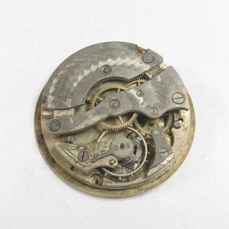 Puede incluir: Primer plano de un movimiento de reloj vintage con engranajes y componentes met&aacute;licos visibles. El mecanismo del reloj es plateado y dorado, con el texto "2 Jewels Adjustment Eanete Swiss" visible en la placa met&aacute;lica.