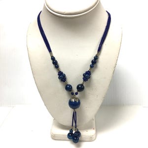 May include: A blue beaded necklace with a pendant. The necklace features a dark blue cord with small beads, larger blue beads, and a central pendant with a large blue orb. The pendant is accented with silver-toned metal and a tassel of blue beads.