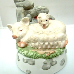 May include: A white porcelain musical piggy bank with a sleeping pig and a piglet on top. The piglet is standing on the sleeping pig's back. The base of the bank is decorated with a green grass and gray stone design.