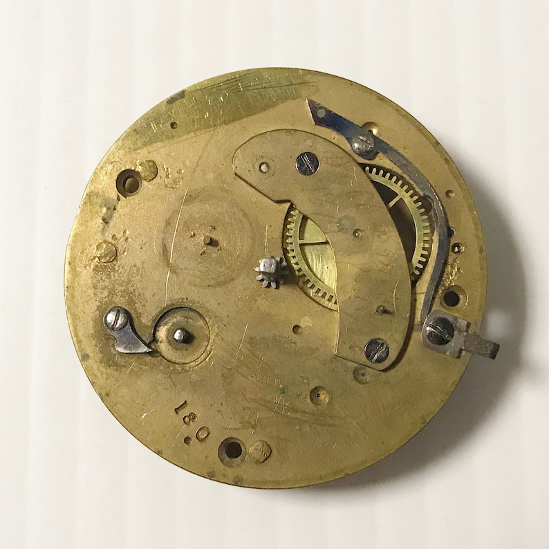 Pu&ograve; includere: Primo piano di un movimento di orologio dorato con ingranaggi visibili e un meccanismo di carica. Il movimento &egrave; contrassegnato dal numero "180."