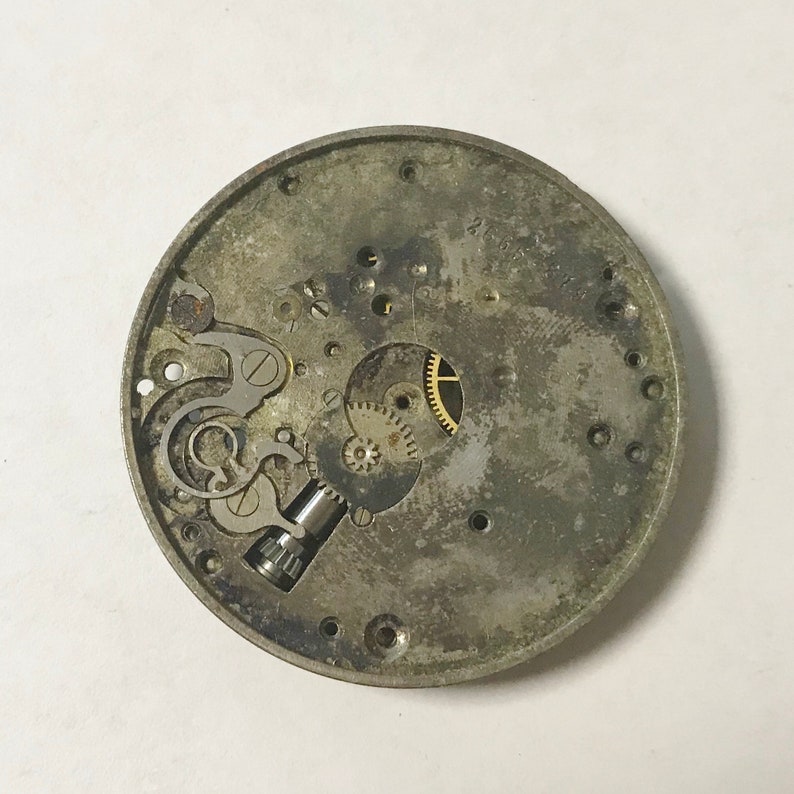 K&ouml;nnte beinhalten: Nahaufnahme eines silbernen Uhrwerks mit freiliegenden Zahnr&auml;dern und einer kleinen Schraube. Das Zifferblatt der Uhr fehlt und die R&uuml;ckseite ist aus Metall.