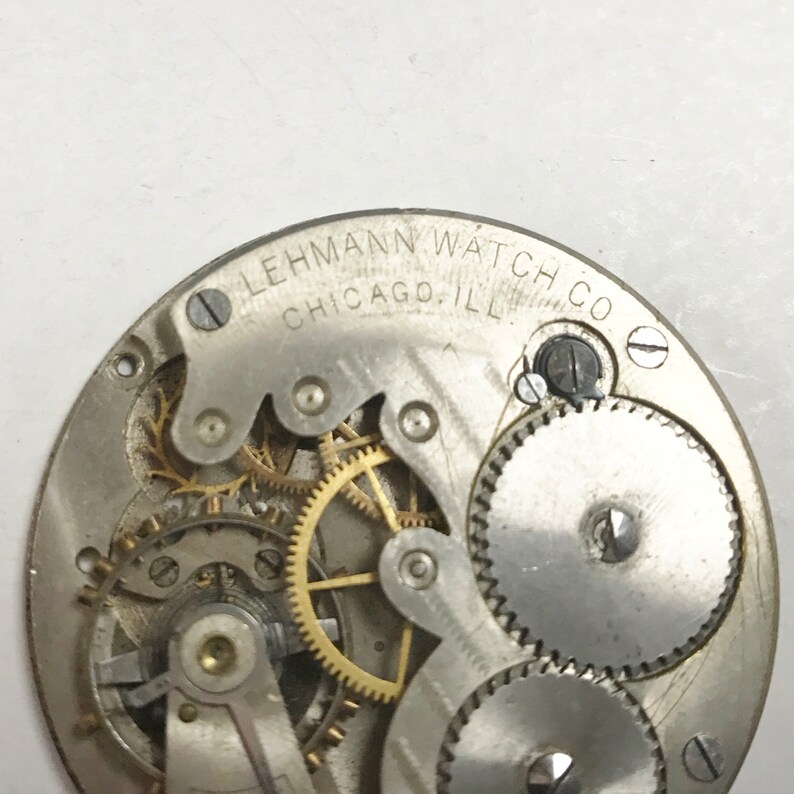 以下が含まれることがあります： 「LEHMANN WATCH CO. CHICAGO. ILL.」という刻印がされた銀色の時計のムーブメントのクローズアップ
