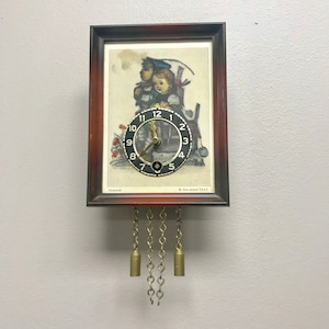 May include: A vintage wooden cuckoo clock with a black face and gold hands. The clock features a picture of two children in traditional German clothing. The clock face has the text "Hummel" and "Ave Maria 9665".