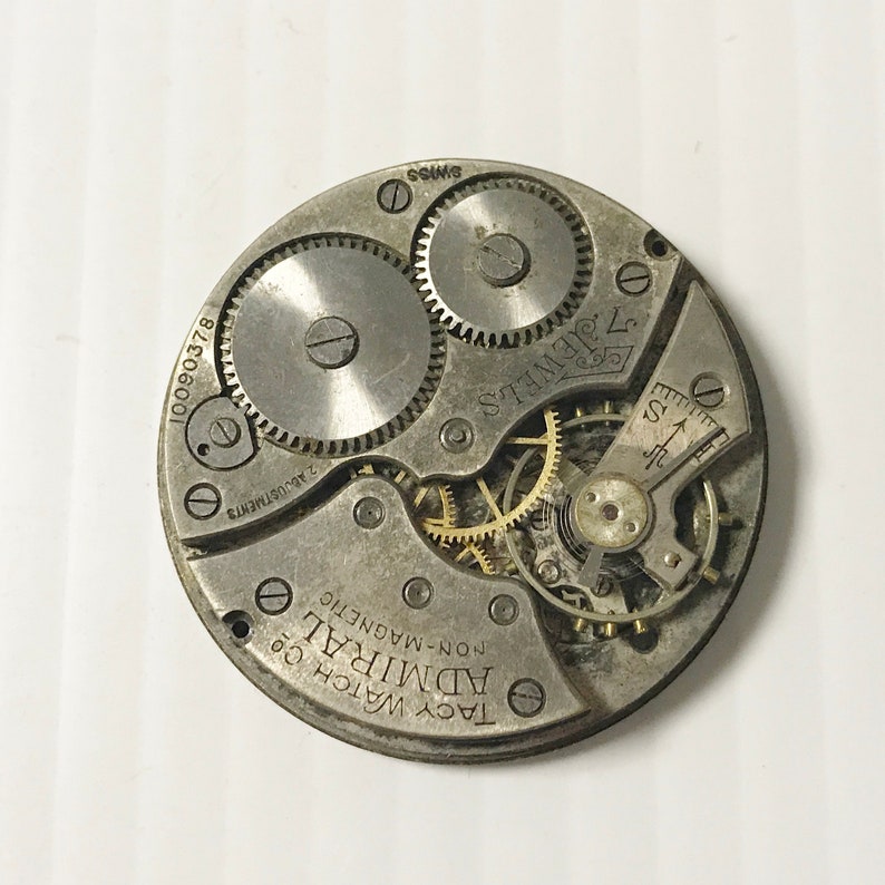 May include: Close-up of a silver pocket watch movement with the text "Tacy Watch Co. Admiral Non-Magnetic 17 Jewels 2 Adjustments 10090378 Swiss" printed on the face.