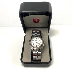 May include: A silver-toned wristwatch with a white face, black numerals, and a stainless steel band is displayed in a black box. The watch face features the number 12 at the top and the brand name. The box has a red and white emblem.