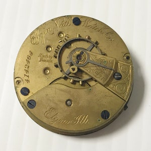May include: Close-up of the back of a gold-colored pocket watch with the inscription "Elgin National Watch Co." and "Elgin Ill." The watch has a safety pinion and the serial number 4142462.