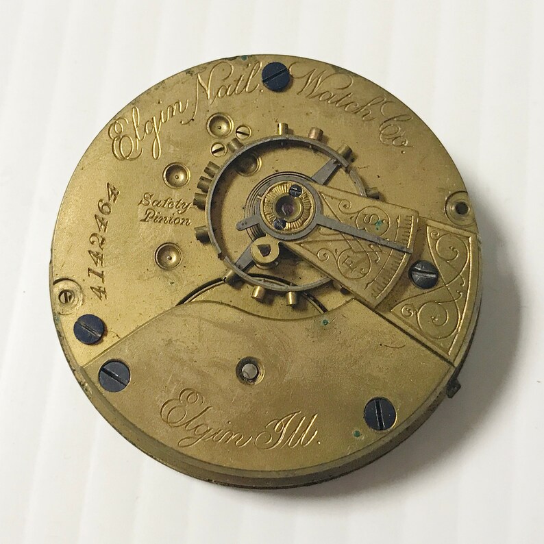 May include: Close-up of the back of a gold-colored pocket watch with the inscription "Elgin National Watch Co." and "Elgin Ill." The watch has a safety pinion and the serial number 4142462.