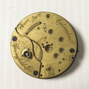 May include: A close-up of a vintage watch movement with a brass finish. The watch mechanism features the text "Springfield Ill." and "Columbia" in cursive script. The serial number "No. 199649" is also visible.