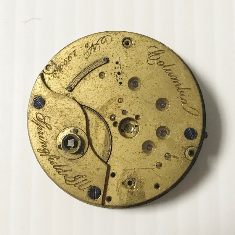 May include: A close-up of a vintage watch movement with a brass finish. The watch mechanism features the text "Springfield Ill." and "Columbia" in cursive script. The serial number "No. 199649" is also visible.