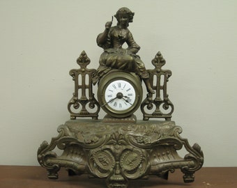 May include: A vintage brass clock with a figurine of a woman sitting on top. The clock face is white with black Roman numeral hour markers and black hands. The clock is decorated with ornate details.