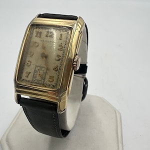 May include: A gold-toned wristwatch with a rectangular face and black leather band. The watch face has Roman numeral hour markers and a small sub-dial at the 6 o'clock position. The watch is on a white surface.