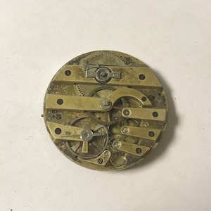 May include: Close-up of a gold-toned pocket watch movement with intricate gears and levers. The watch is disassembled and the inner workings are visible.