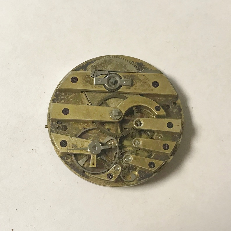 May include: Close-up of a gold-toned pocket watch movement with intricate gears and levers. The watch is disassembled and the inner workings are visible.