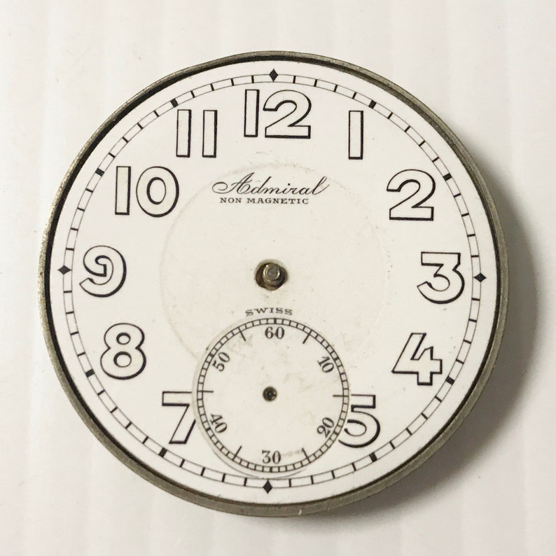 May include: White watch face with black Roman numeral hour markers and black minute markers. The watch face also has a smaller, concentric circle with black markings for seconds. The watch face has the text "Admiral NON MAGNETIC" and "SWISS" printed on it.