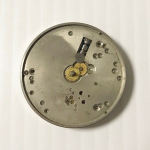 May include: A silver-colored watch movement with visible gears, screws, and other internal components. The number "121377" is stamped on the surface. This item is likely a vintage or antique watch part.