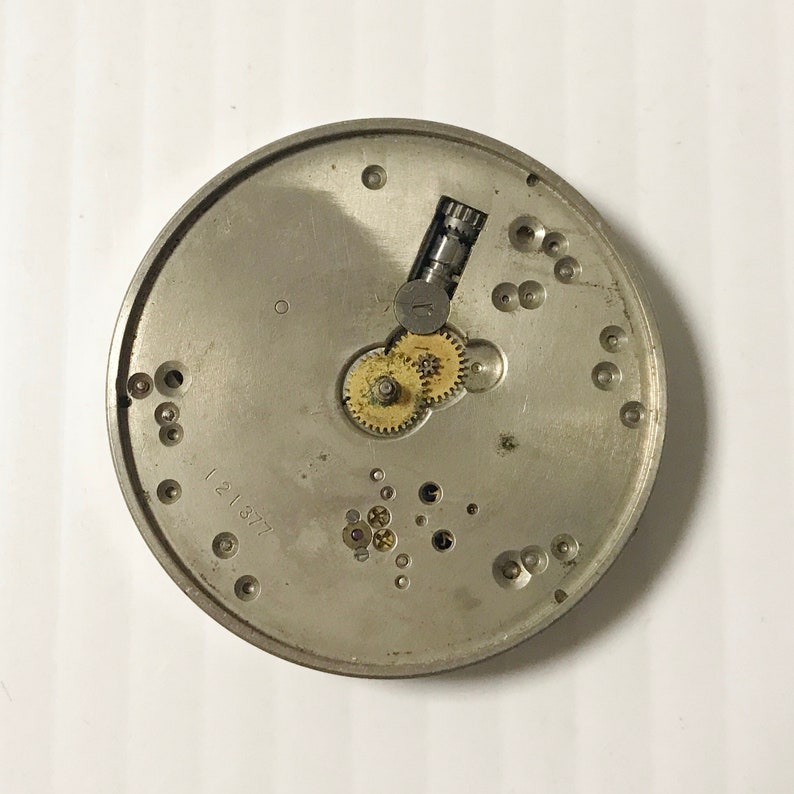 May include: A silver-colored watch movement with visible gears, screws, and other internal components. The number "121377" is stamped on the surface. This item is likely a vintage or antique watch part.
