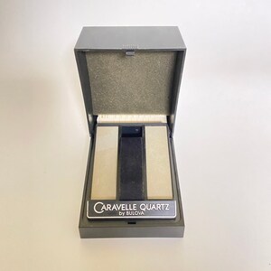 May include: A black watch box with a gray interior and a black label that reads "Caravelle Quartz by Bulova".
