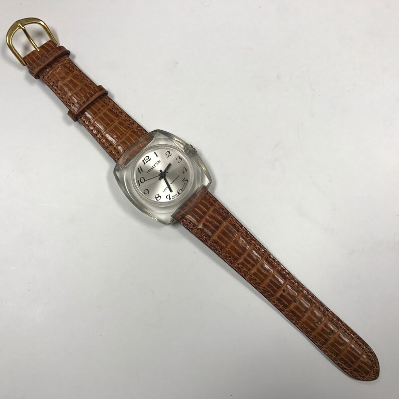 Peut inclure: Une montre-bracelet en plastique transparent avec un bracelet en cuir marron. Le cadran de la montre a des chiffres romains noirs et une aiguille des secondes argent&eacute;e.