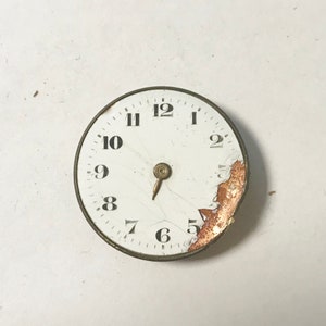May include: A white clock face with black numbers 1 through 12. The clock face is cracked and missing a portion of the outer edge. The clock face is surrounded by a gold-colored metal rim.