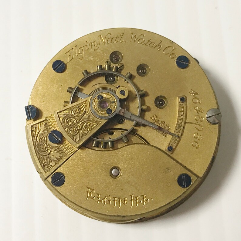 May include: Close-up of a gold-toned pocket watch movement with the inscription "Elgin Natl. Watch Co." and "808046" on the face. The watch has a safety winding mechanism and intricate engravings.
