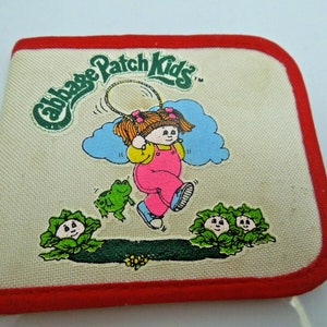 May include: A white canvas Cabbage Patch Kids wallet with a red border. The wallet features a cartoon image of a Cabbage Patch Kid girl jumping rope with two frogs and two cabbage patch kids in the background. The text "Cabbage Patch Kids" is printed in green on the wallet.