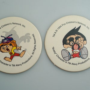 May include: Two round, white, cardboard tokens featuring a cartoon character named "Bobby". The first token shows Bobby wearing a blue hat and running. The second token shows Bobby wearing a red shirt and black glasses. Both tokens have the text "TM & © 1994 Fox Children's Network, Inc. "Bobby" Character is TM Alevy Productions. All Rights Reserved."