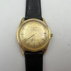 May include: A gold-tone wristwatch with a black leather band. The watch face is white with black markings and the text "Eterna Matic" and "Chronometre".