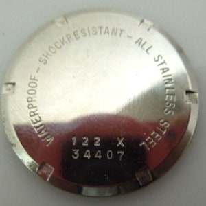 May include: The back of a silver watch with the text "Waterproof - Shockresistant - All Stainless Steel" and the numbers "122 x 344 07".