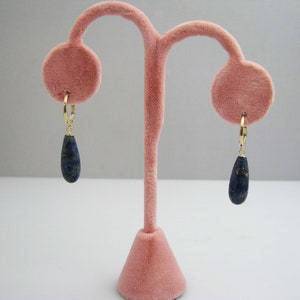 May include: A pair of gold hoop earrings with teardrop-shaped lapis lazuli stones hanging from them. The earrings are displayed on a pink velvet stand.