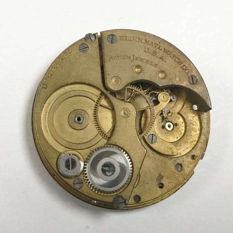 K&ouml;nnte beinhalten: Nahaufnahme eines goldfarbenen Taschenuhrwerks mit der Aufschrift "ELGIN NATL WATCH CO. U.S.A. SEVENTEEN JEWELS" und der Nummer "8388391".