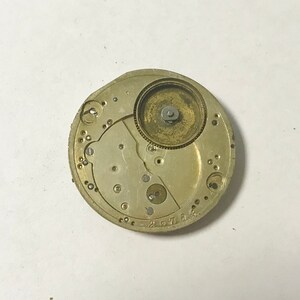 May include: Close-up of a gold-toned watch movement with intricate gears and a circular winding mechanism. The movement is set against a white background.